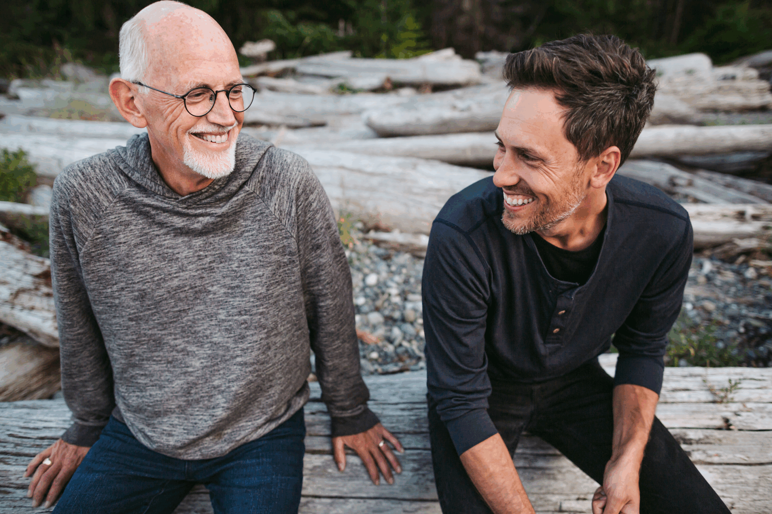 And elderly man and middle aged gentleman sat on a log in the forest smiling at each other